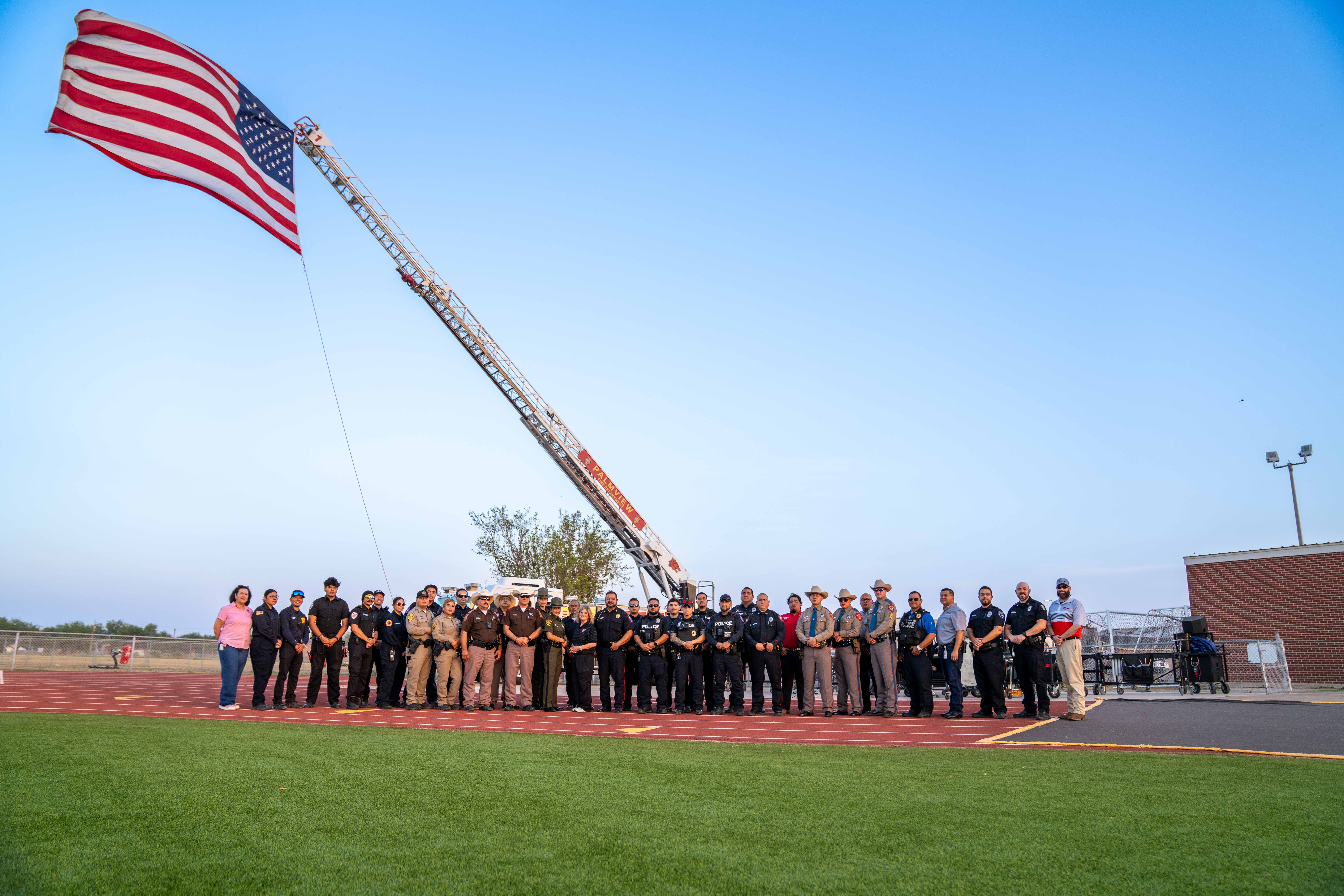 A large group of diverse first responders poses outdoors on a running track with a raised American flag.