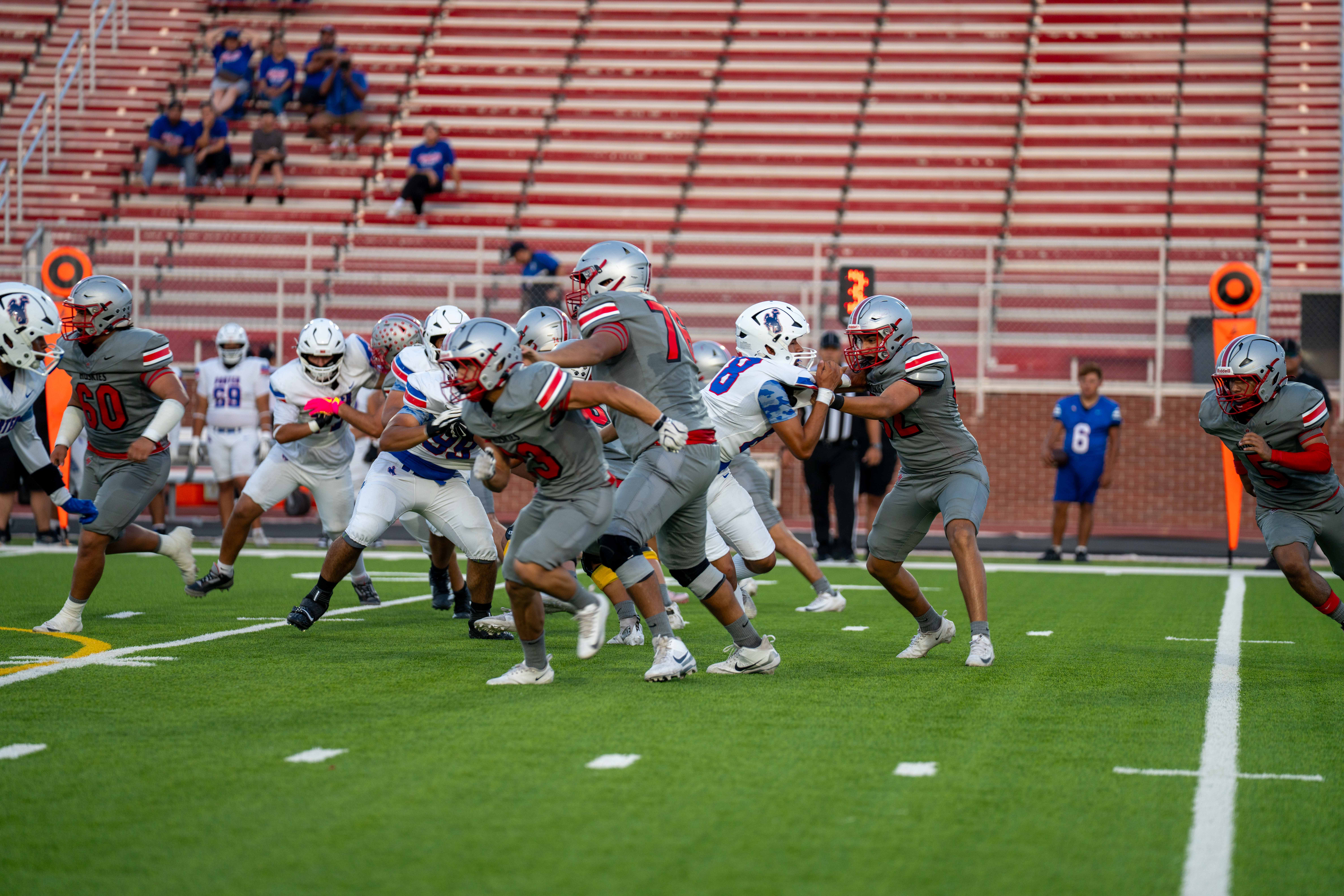 A high school football game is underway on a green turf field with players in gray and blue uniforms.