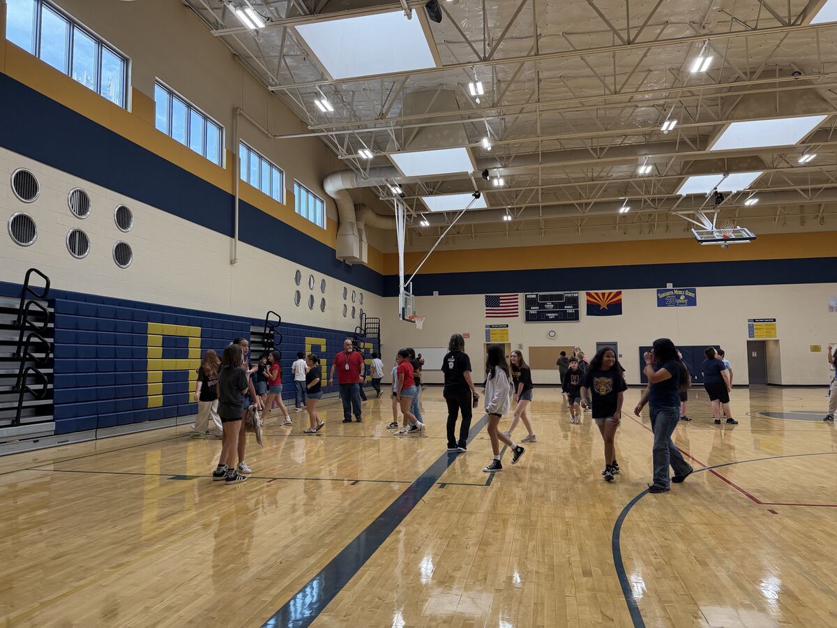 Students and adults gather on a polished wooden gymnasium floor, with bleachers and a basketball hoop visible.