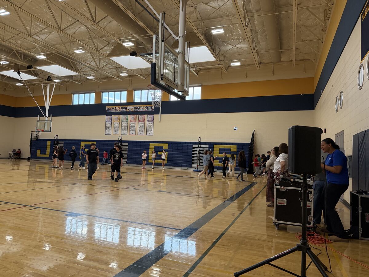 Students and adults gather in a school gymnasium with a basketball hoop and bleachers.