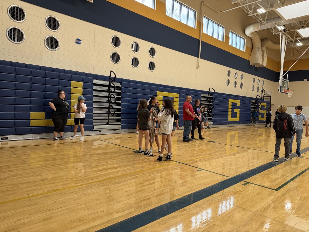 A group of people, mostly young students, stand on a polished wooden gymnasium floor.