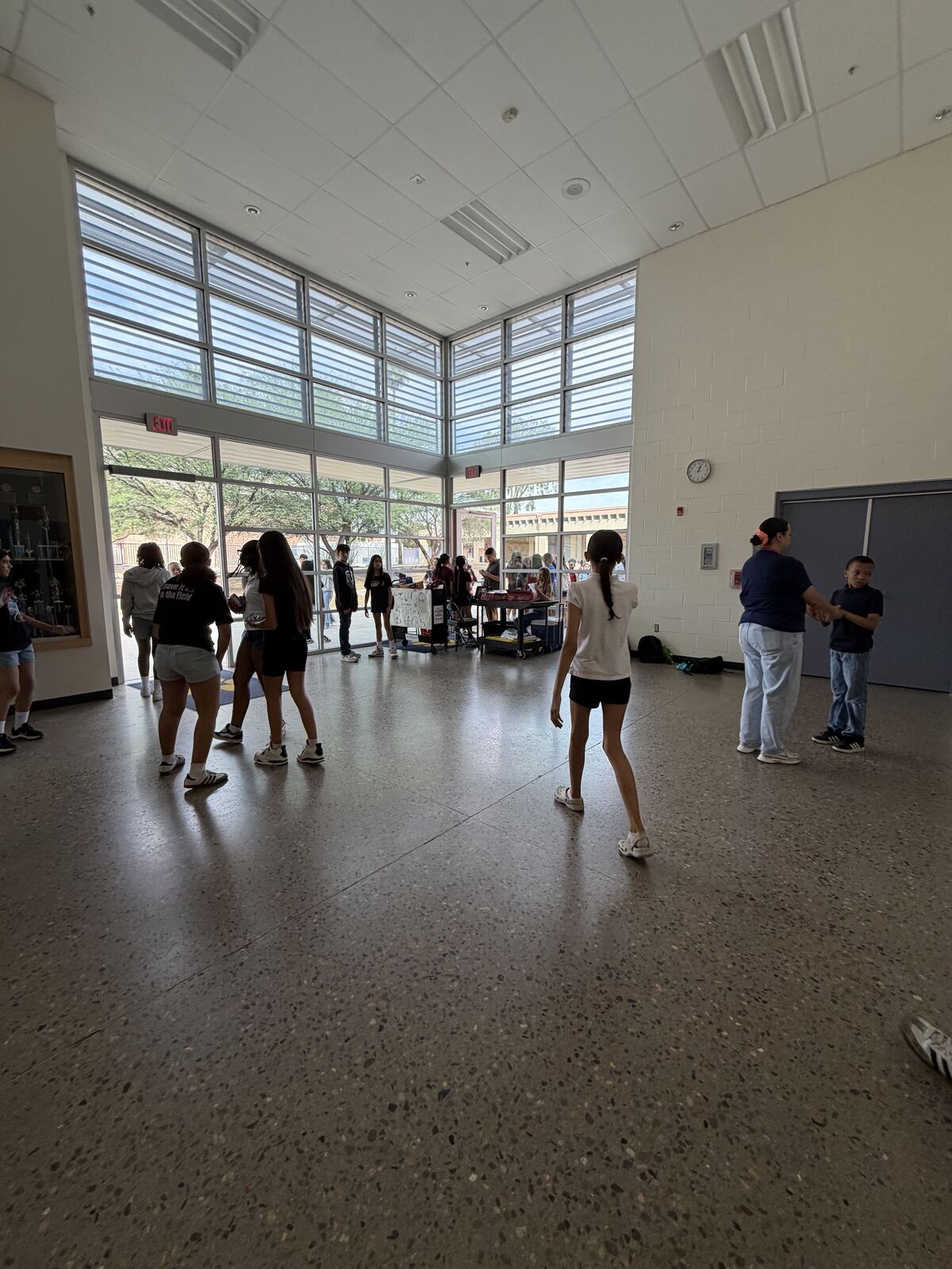 A group of students stand in a line in a hallway, facing large windows.