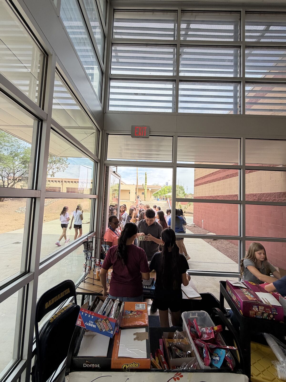 A group of people are gathered around tables laden with snacks and boxes, possibly at a school event or fundraiser.