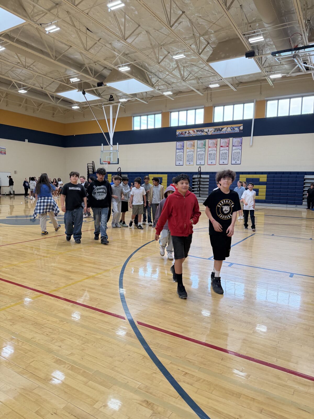 Students stand in a line on a polished wooden gymnasium floor, with a basketball hoop visible in the background.