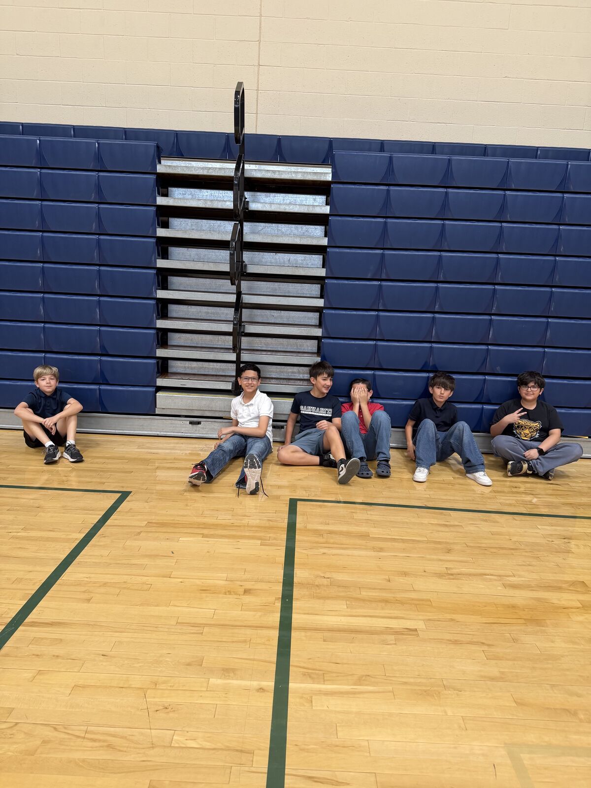 A group of young people sit on the floor of a gymnasium next to bleachers.