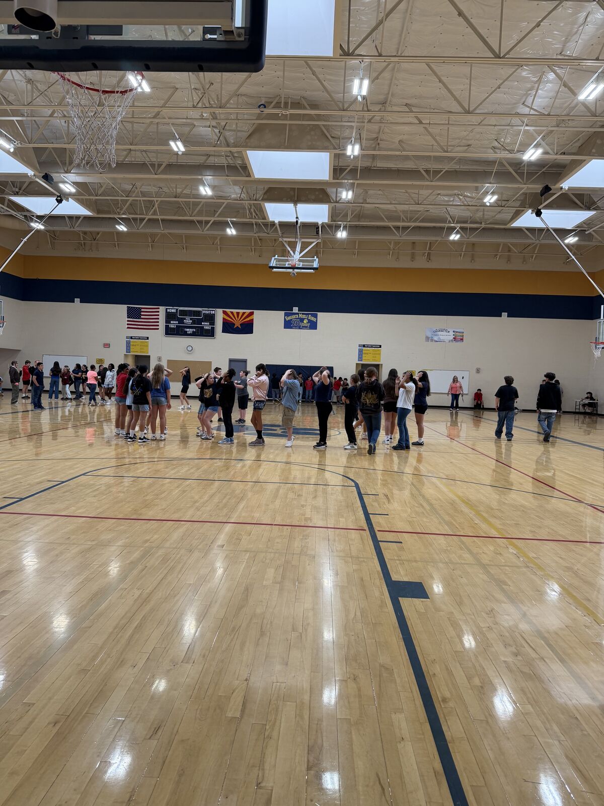 A group of students stand in a line on a polished wooden gymnasium floor, facing a wall with banners and a scoreboard.