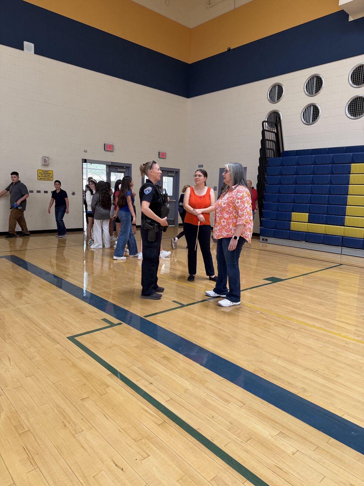 An SRO stands in a gymnasium, talking to teachers surrounded by a group of students.