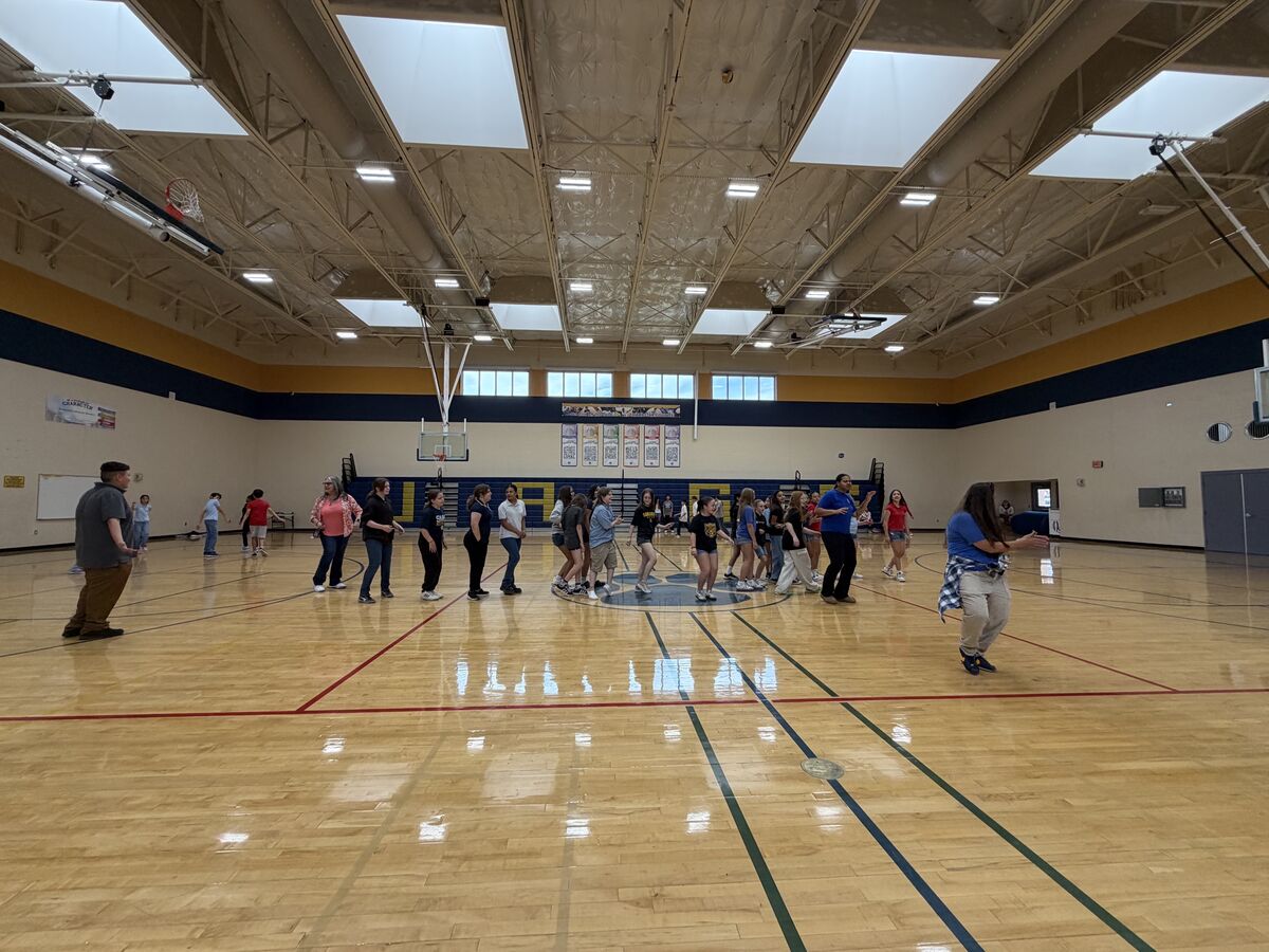 A group of students and adults are gathered on a polished wooden gymnasium floor.