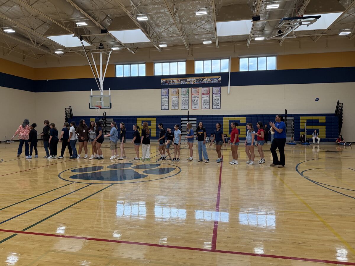 A group of students and an adult stand in a line on a polished gymnasium floor, with a large paw print logo visible.