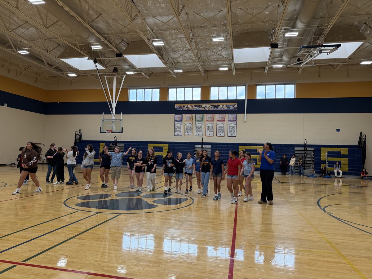 A group of students stand on a basketball court, some looking towards a woman in a blue shirt.