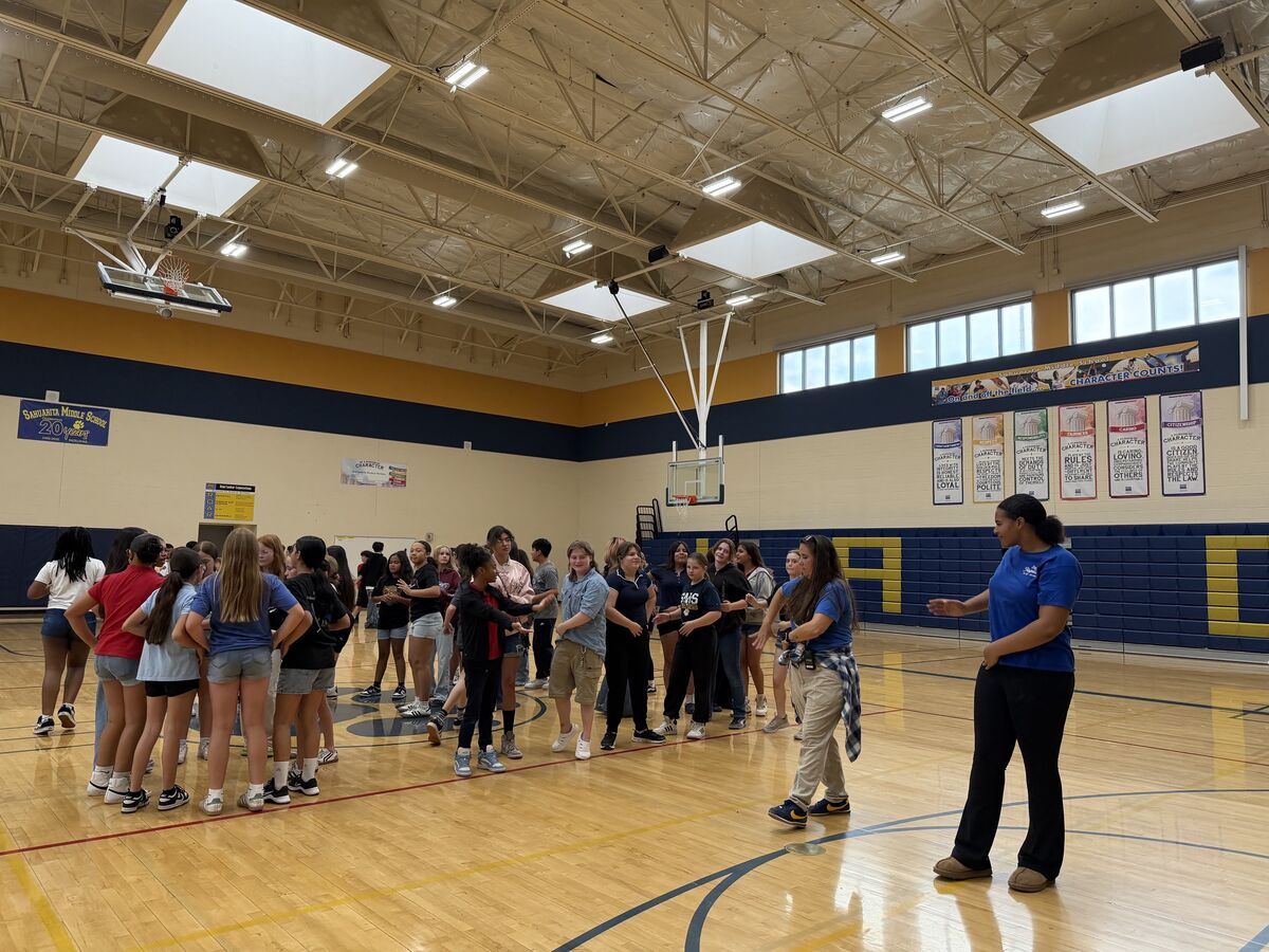 A group of students and adults gather in a school gymnasium, with banners displaying 'CHARACTER COUNTS!' visible on the wall.