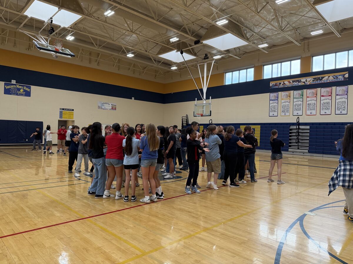 A group of middle school students stand in a circle on a polished gymnasium floor, facing each other.
