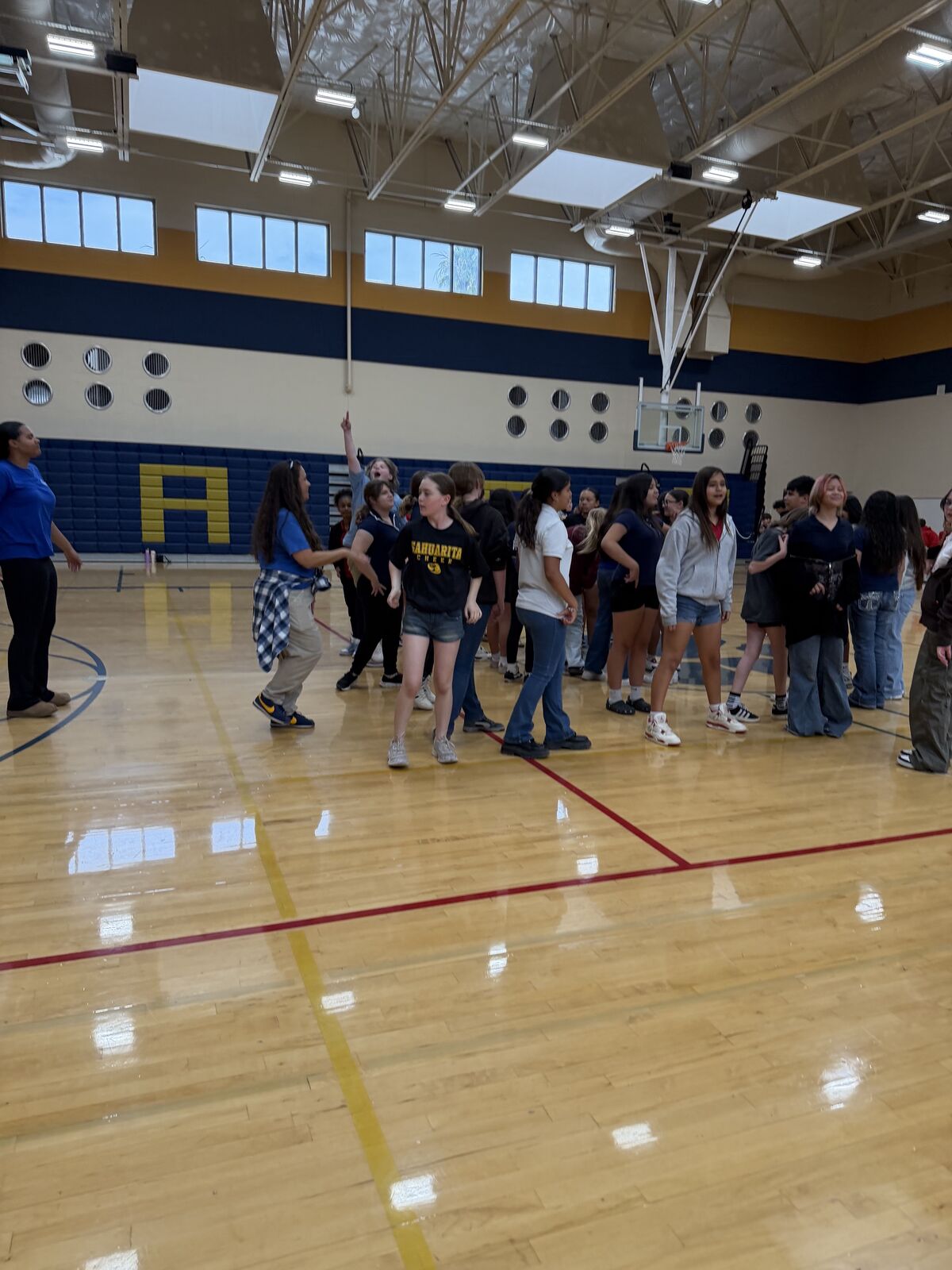 A group of young people stand on a polished wooden gym floor, some looking towards the camera.
