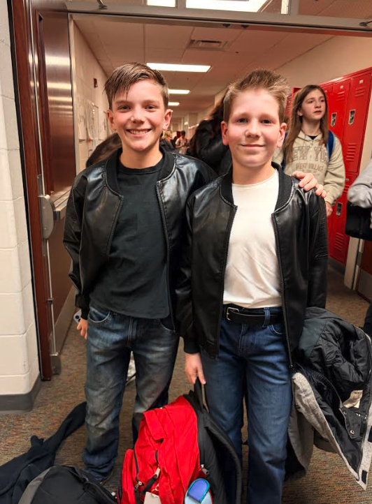 Two smiling boys in black leather jackets and jeans stand in a school hallway.