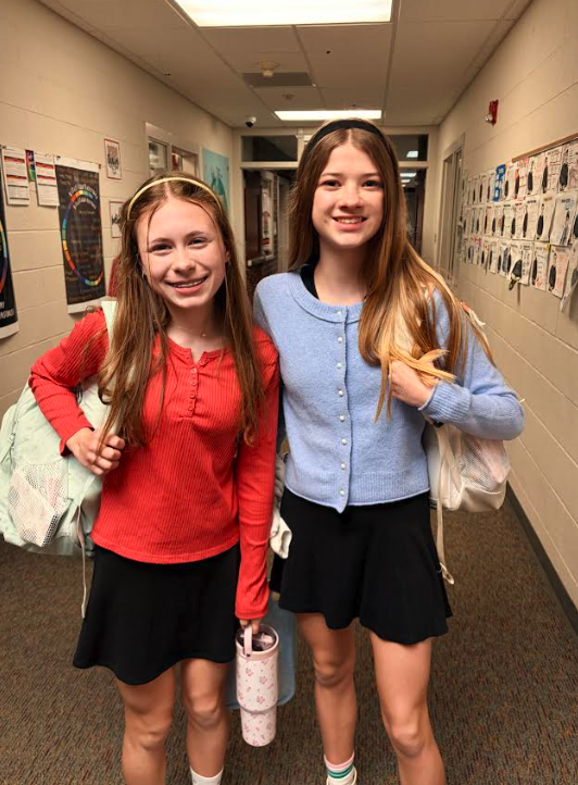 Two smiling young girls stand in a school hallway, each wearing black skirt and cardigan.