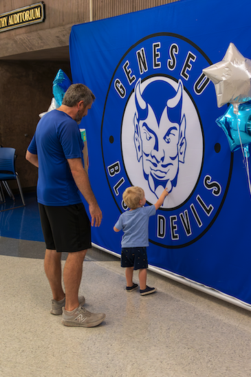 A man and a young child stand before a large blue banner that reads "GENESEO BLUE DEVILS" with a mascot logo.