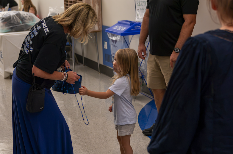 A woman in a blue skirt hands blue bead necklaces to a young girl indoors.