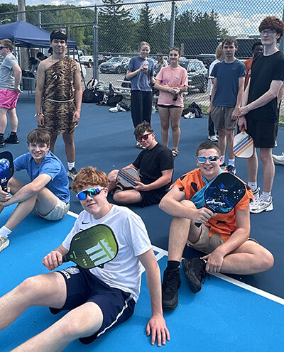 A group of young people in casual attire and costumes sit on a blue court, holding pickleball paddles.