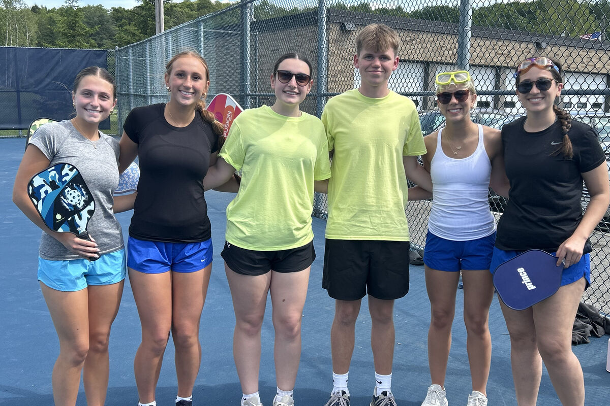 Six young people in athletic wear stand on a blue court, holding pickleball paddles.