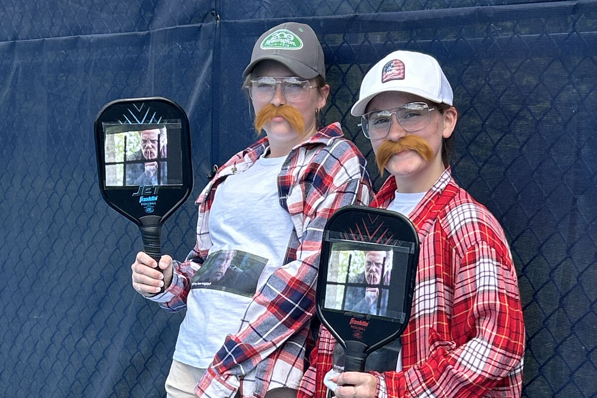 Two people in plaid shirts and fake mustaches hold pickleball paddles with images of a man's face.
