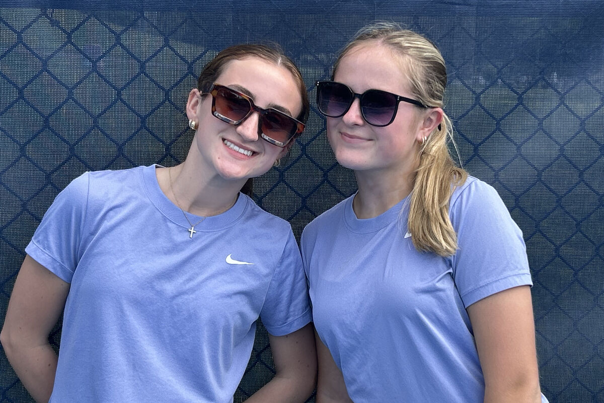 Two young women in matching light blue Nike t-shirts and sunglasses smile at the camera.