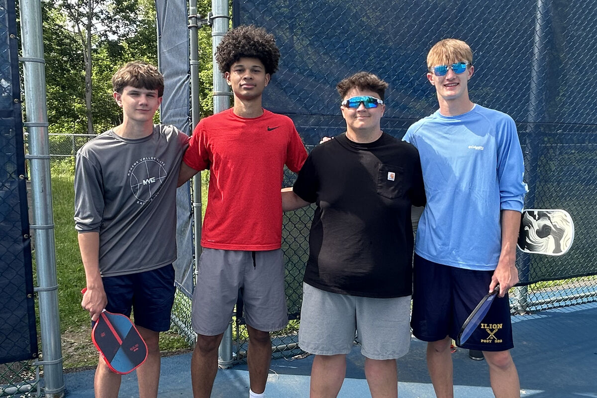 Four young men stand together on a blue court, holding pickleball paddles.
