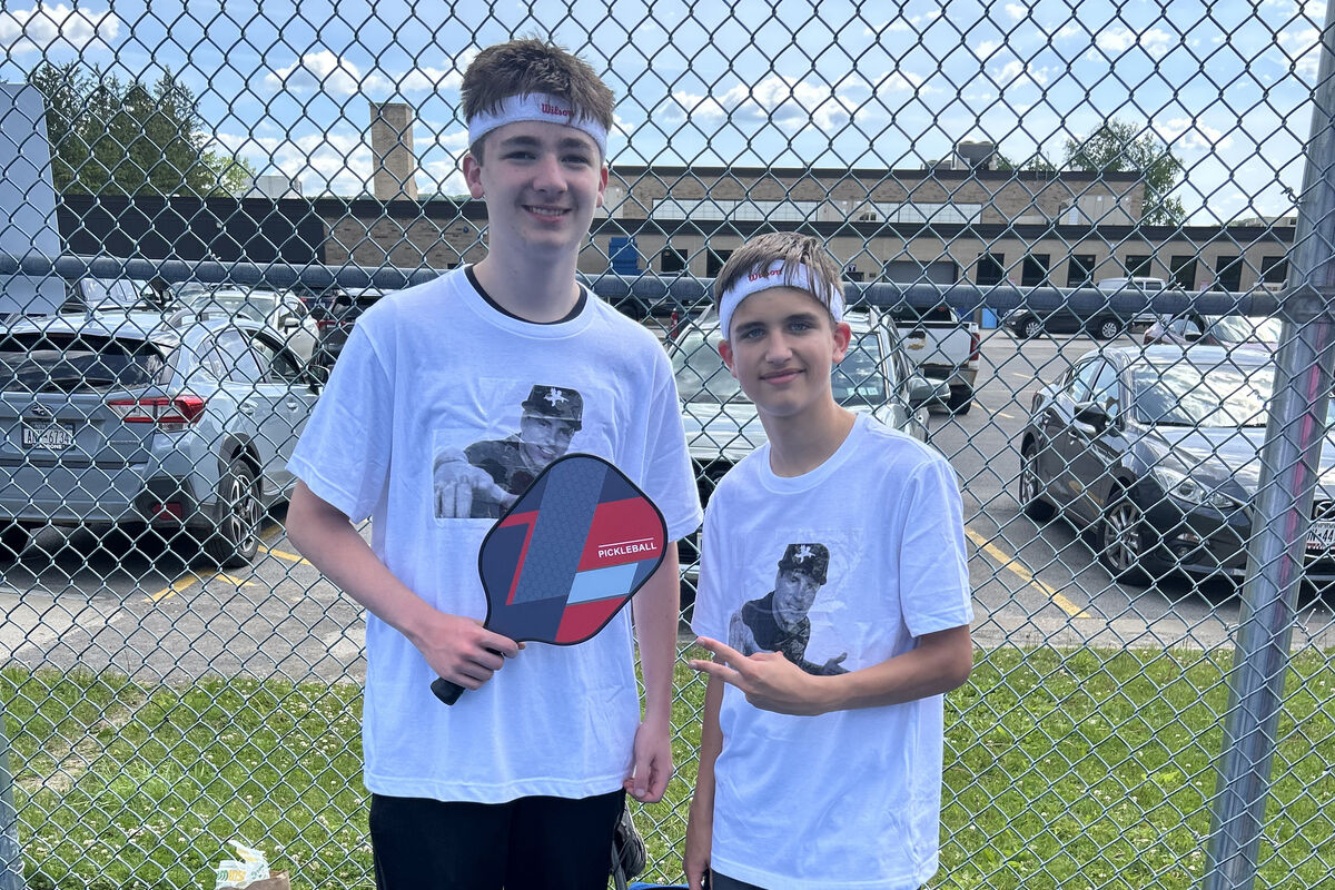 Two smiling young men in matching t-shirts and headbands stand behind a chain-link fence, one holding a pickleball paddle.