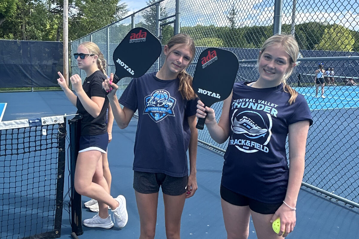Three young women stand on a blue pickleball court, holding pickleball paddles.