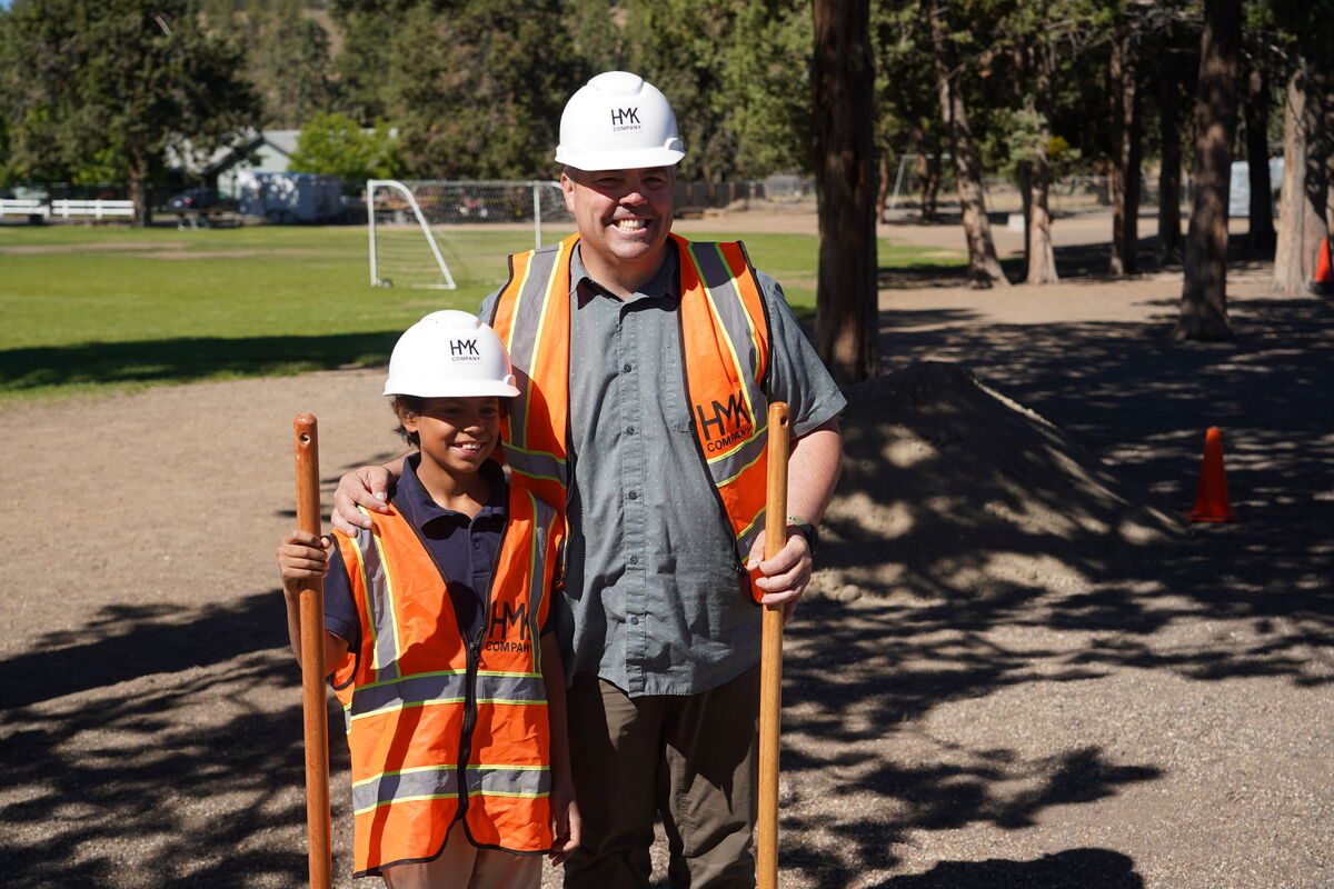 A man and a child, both wearing hard hats and orange safety vests, stand together outdoors.