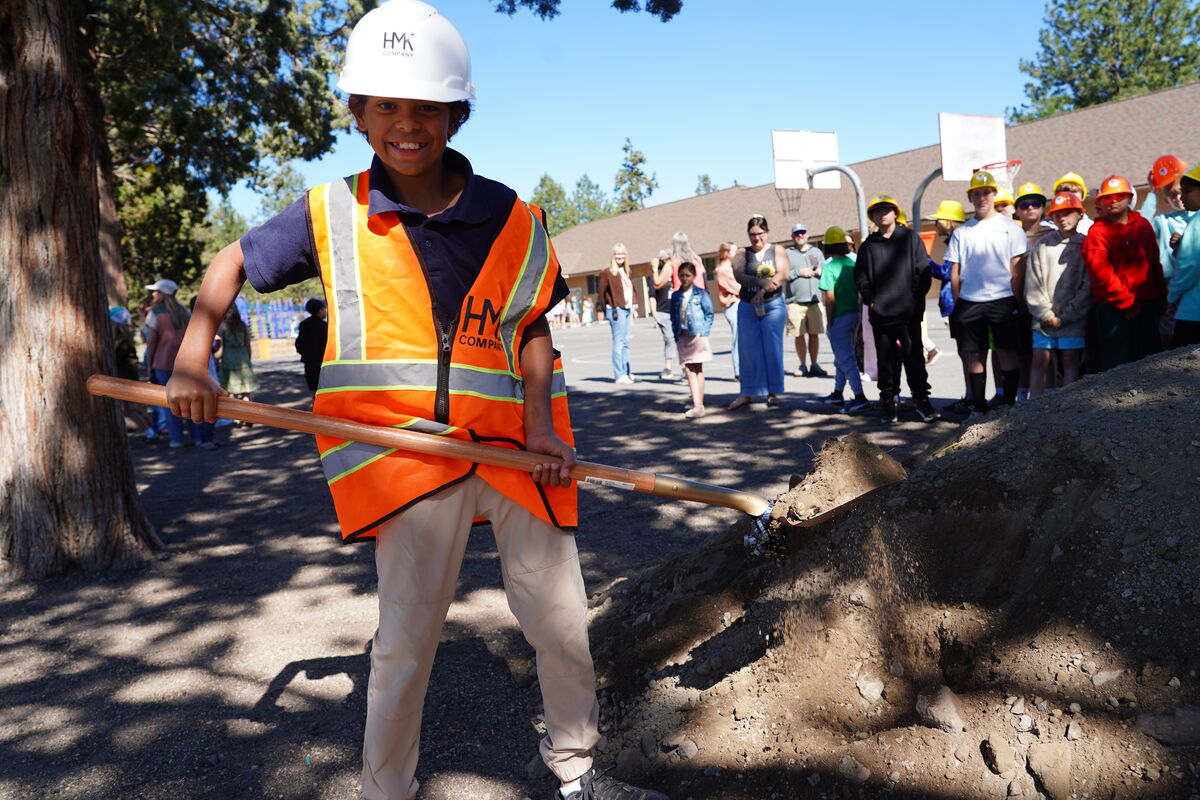 A young boy in a white hard hat and orange safety vest smiles while holding a shovel near a pile of dirt.