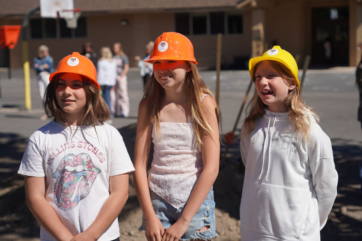Three young girls wearing hard hats stand outdoors, smiling.