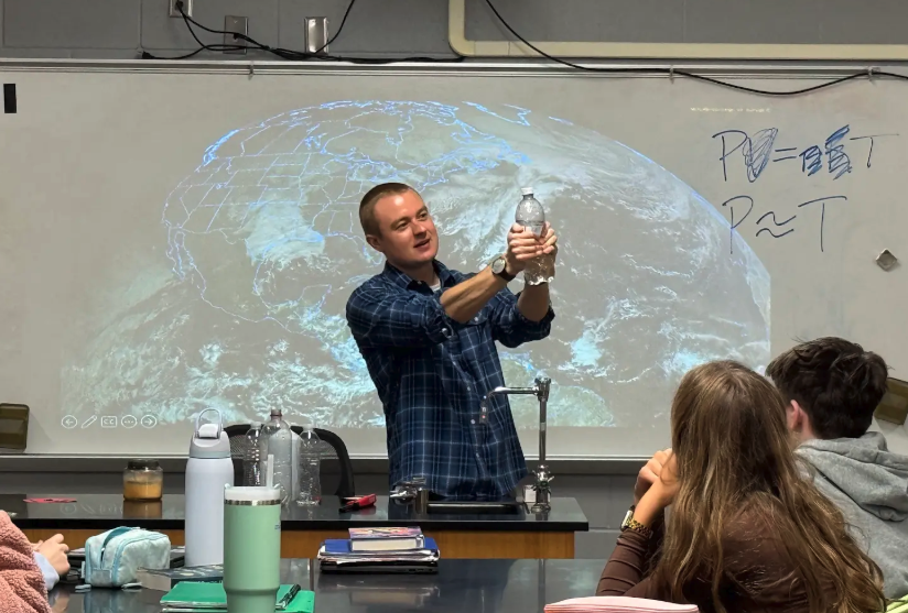 A teacher demonstrates a science experiment with a water bottle in front of a projected image of Earth.