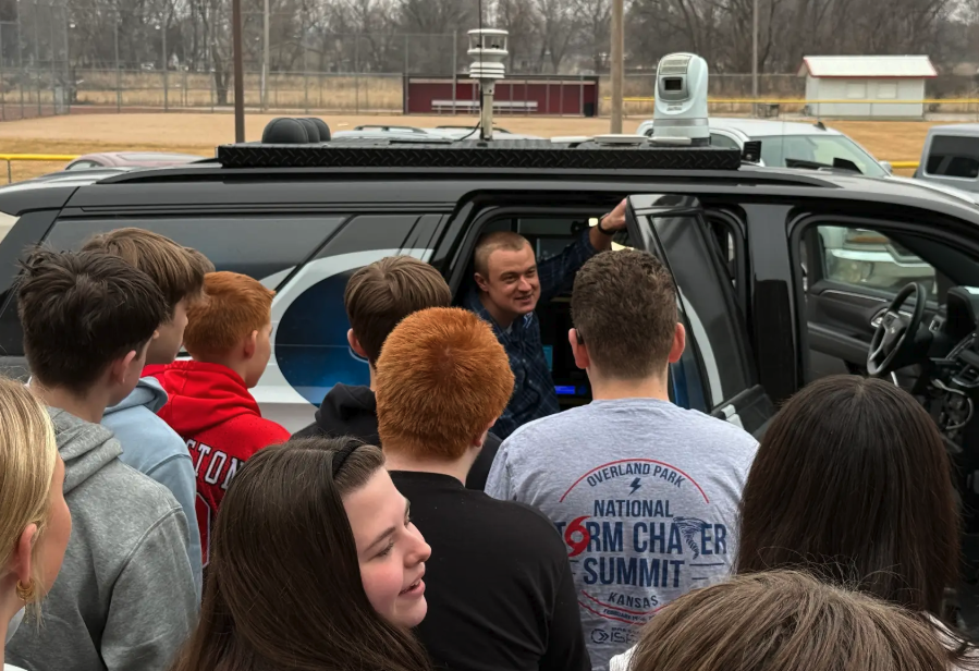A man in a plaid shirt smiles while standing by the open door of a vehicle equipped with weather monitoring equipment.