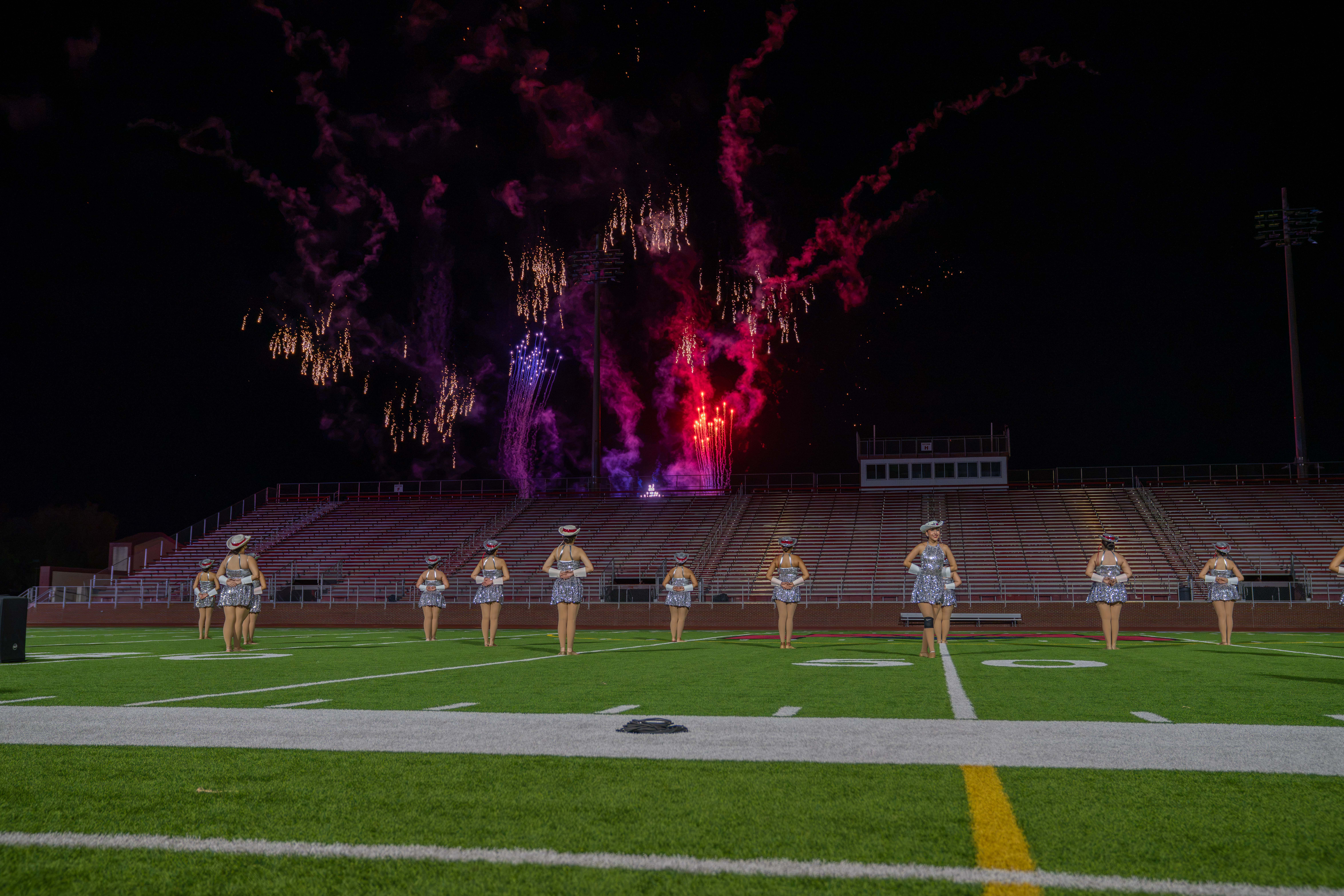 A group of performers in sparkly outfits stand on a football field at night, with fireworks illuminating the sky.