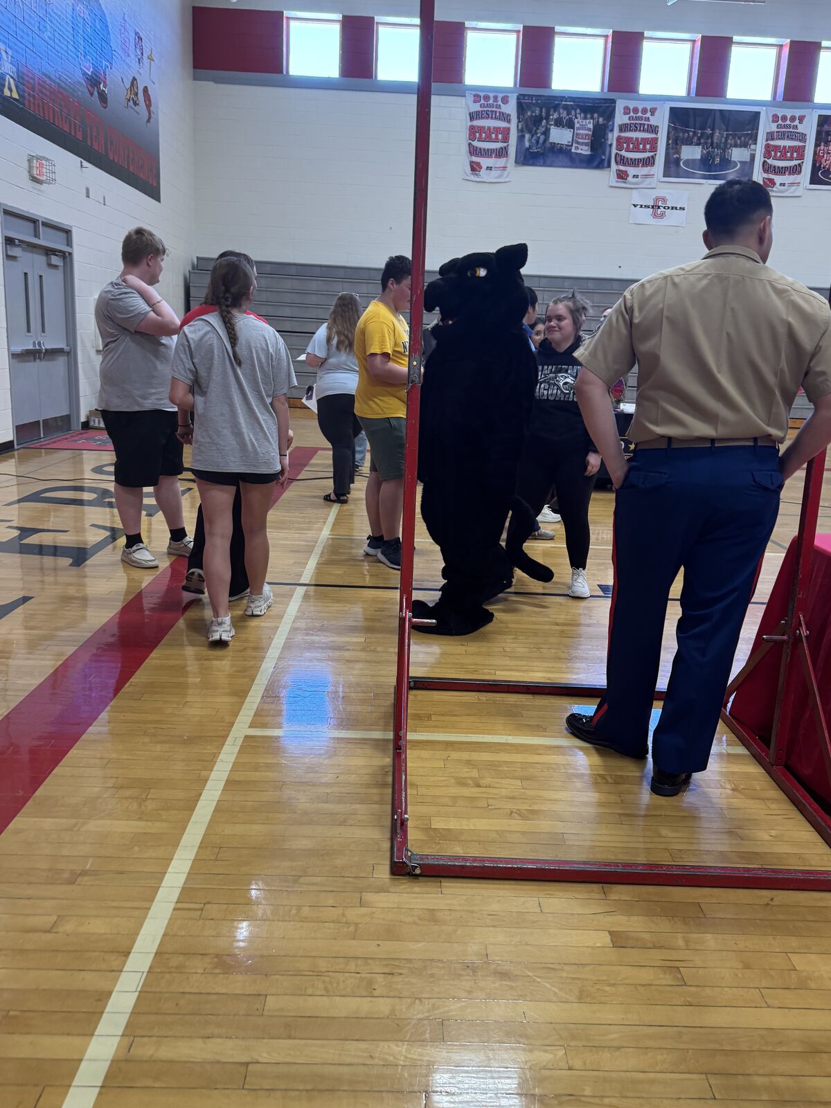 A group of students and a person in a military uniform stand in a gymnasium with wrestling championship banners on the wall.