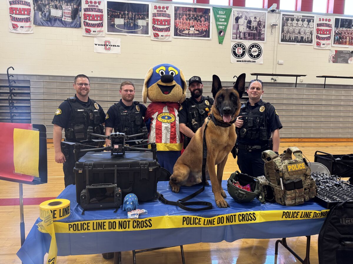 Four police officers, a police dog, and a mascot stand behind a table displaying police equipment, including a drone and tactical gear.