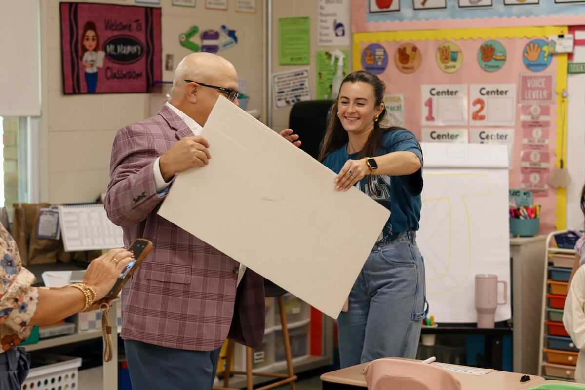 A smiling man and woman hold up a large white poster board in a colorful elementary school classroom.