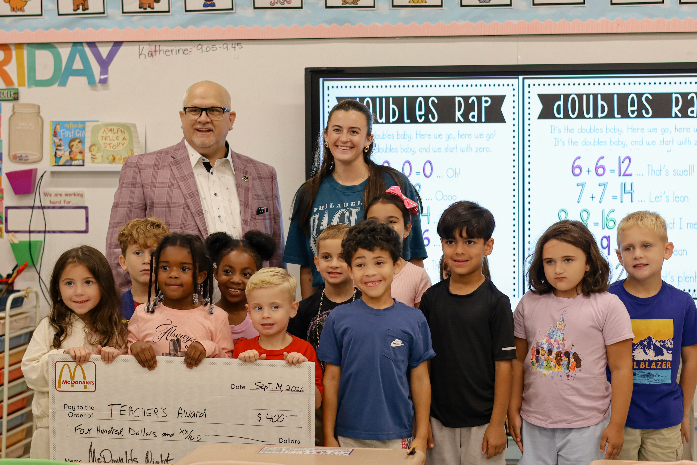A teacher and principal stand with smiling students in a classroom, holding a large novelty check for $400.