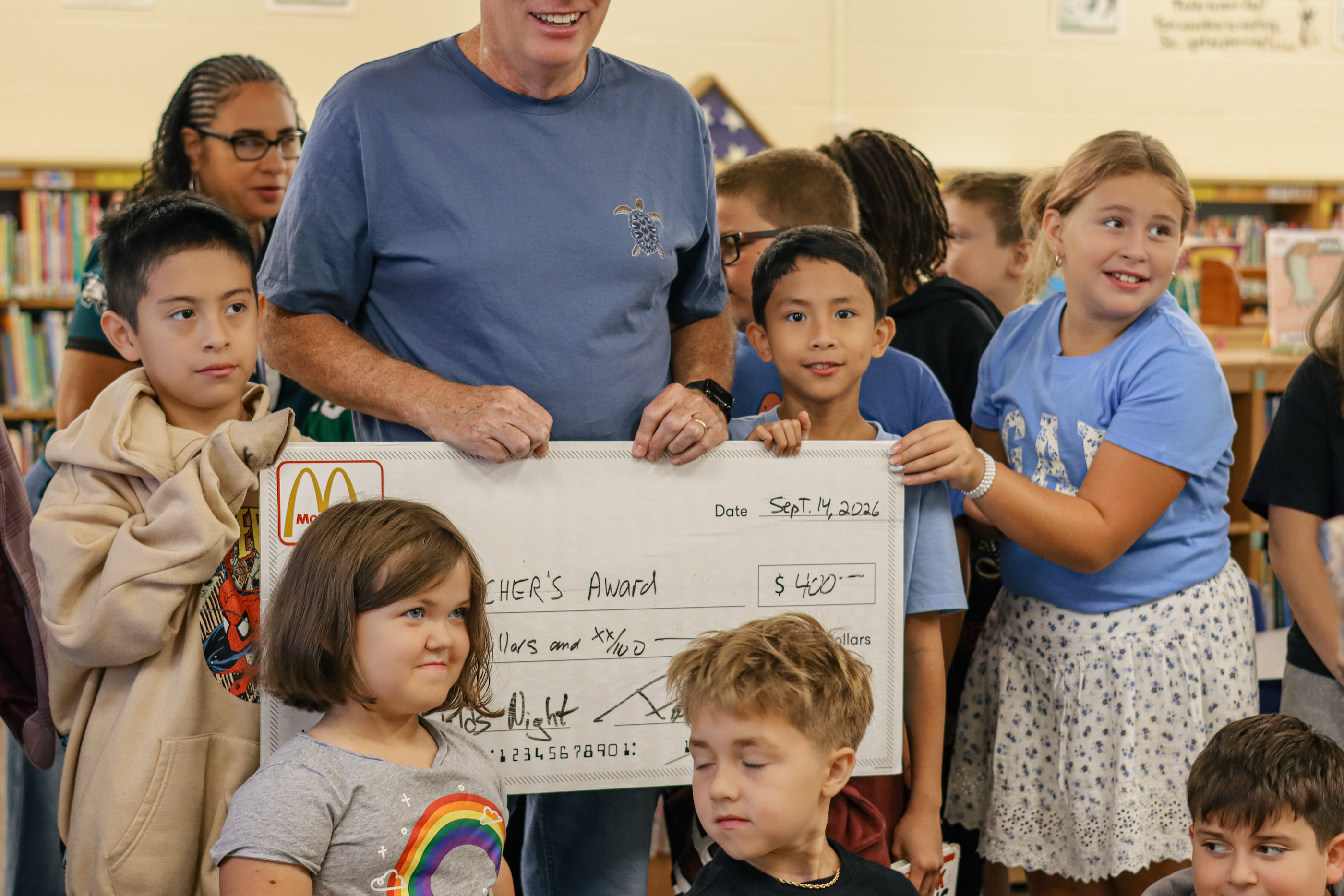 A diverse group of children and a man holding a large novelty check for $400 from McDonald's in a library.