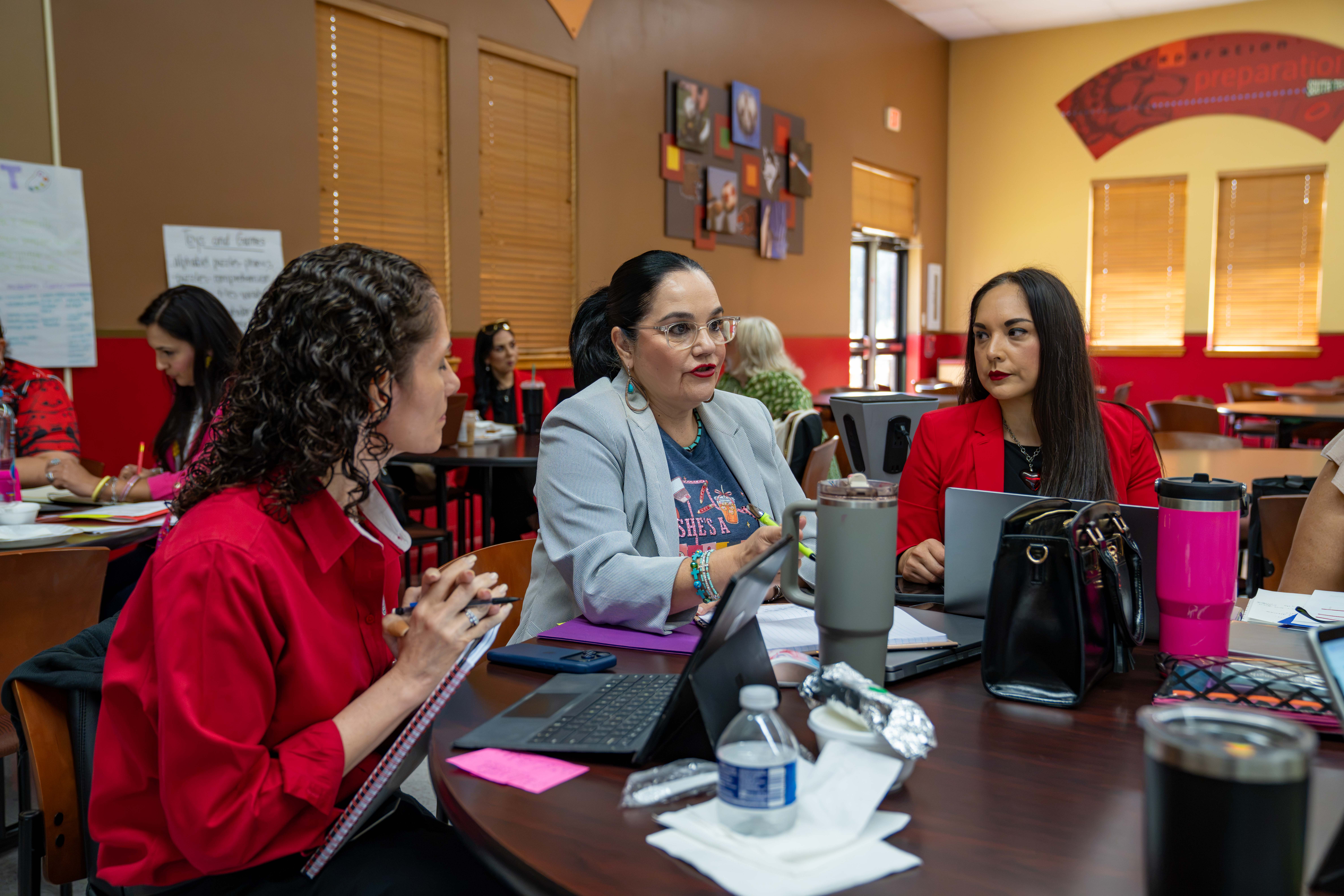 Three women are seated at a table, engaged in a discussion with laptops and notebooks.