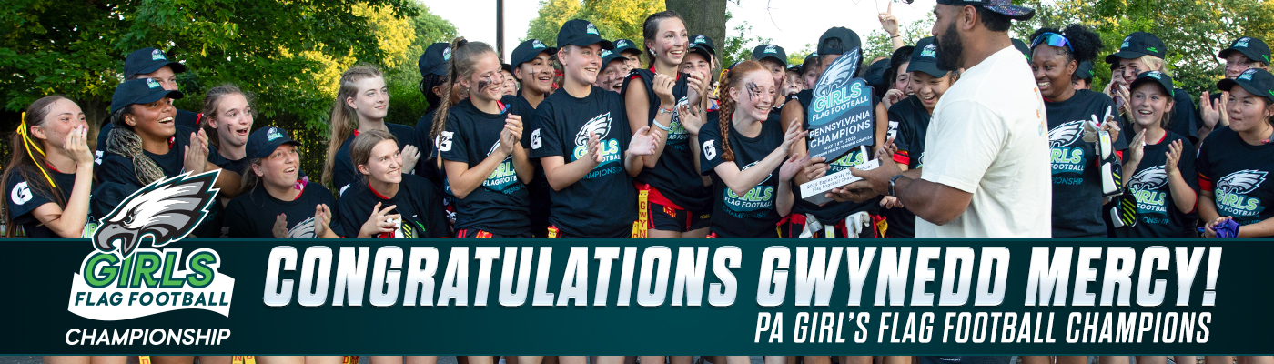 A group of girls in matching black t-shirts and hats celebrate winning a flag football championship, holding a trophy.