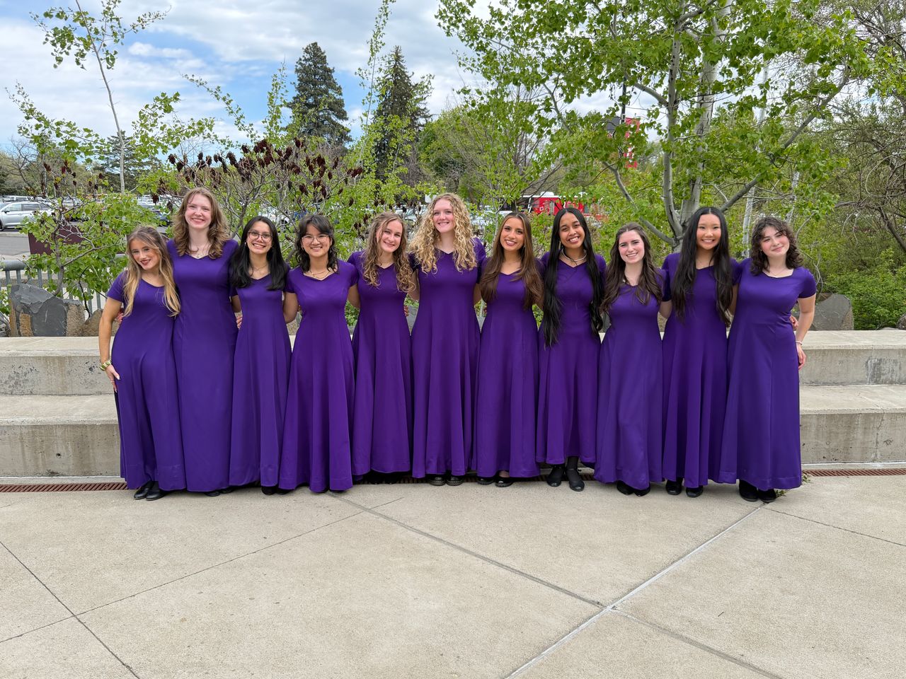 A group of eleven young women in matching purple choir robes stand together outdoors.