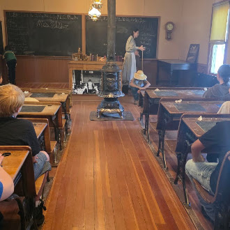 A historic one-room schoolhouse features wooden desks, a central wood-burning stove, and a teacher instructing students.