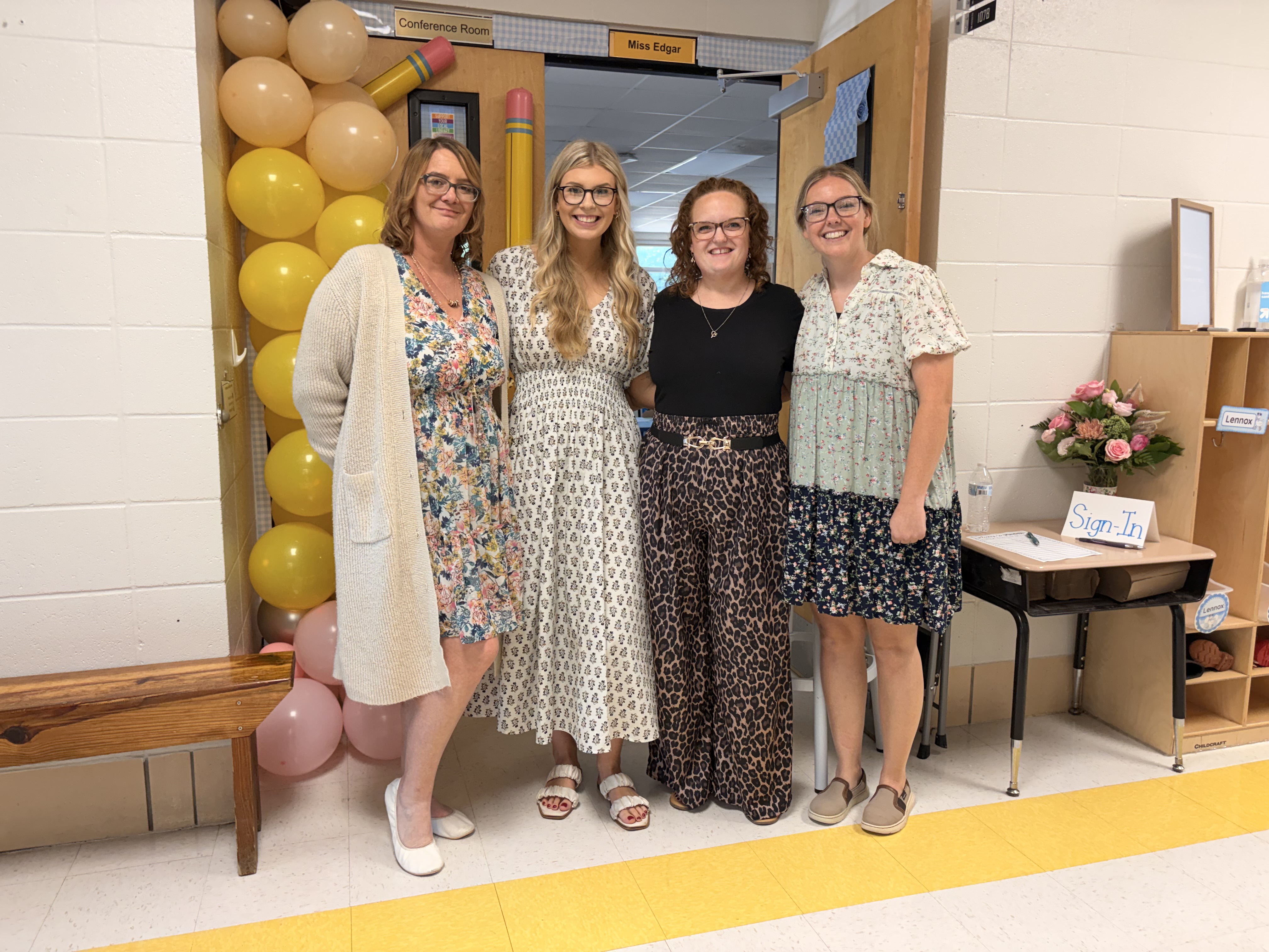 Four women stand together in a hallway decorated with balloons and a large pencil made of balloons.