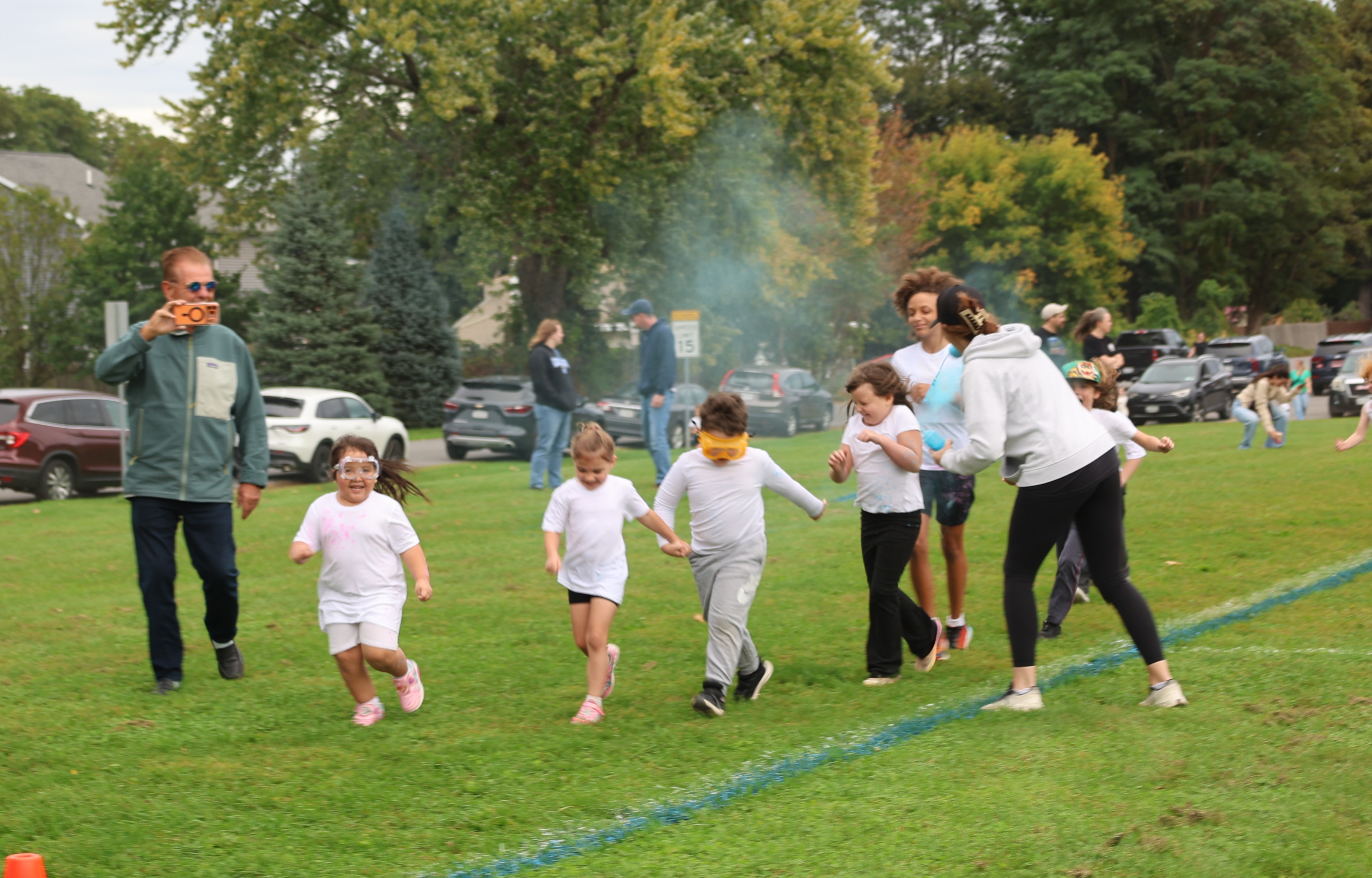 A group of children joyfully run across a grassy field during an outdoor event, with an adult recording video nearby.