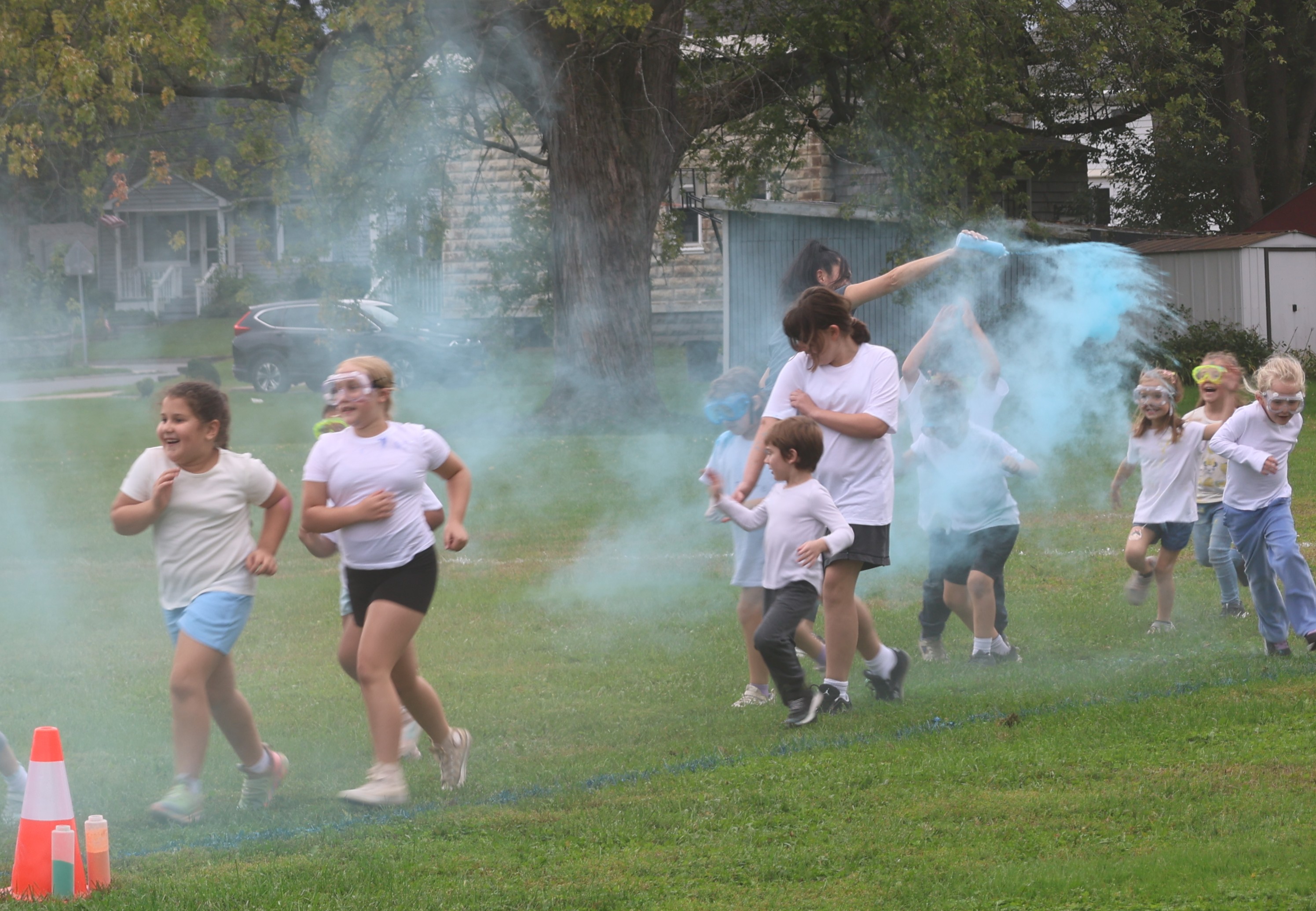 A group of children in white shirts run across a grassy field through a cloud of blue powder.