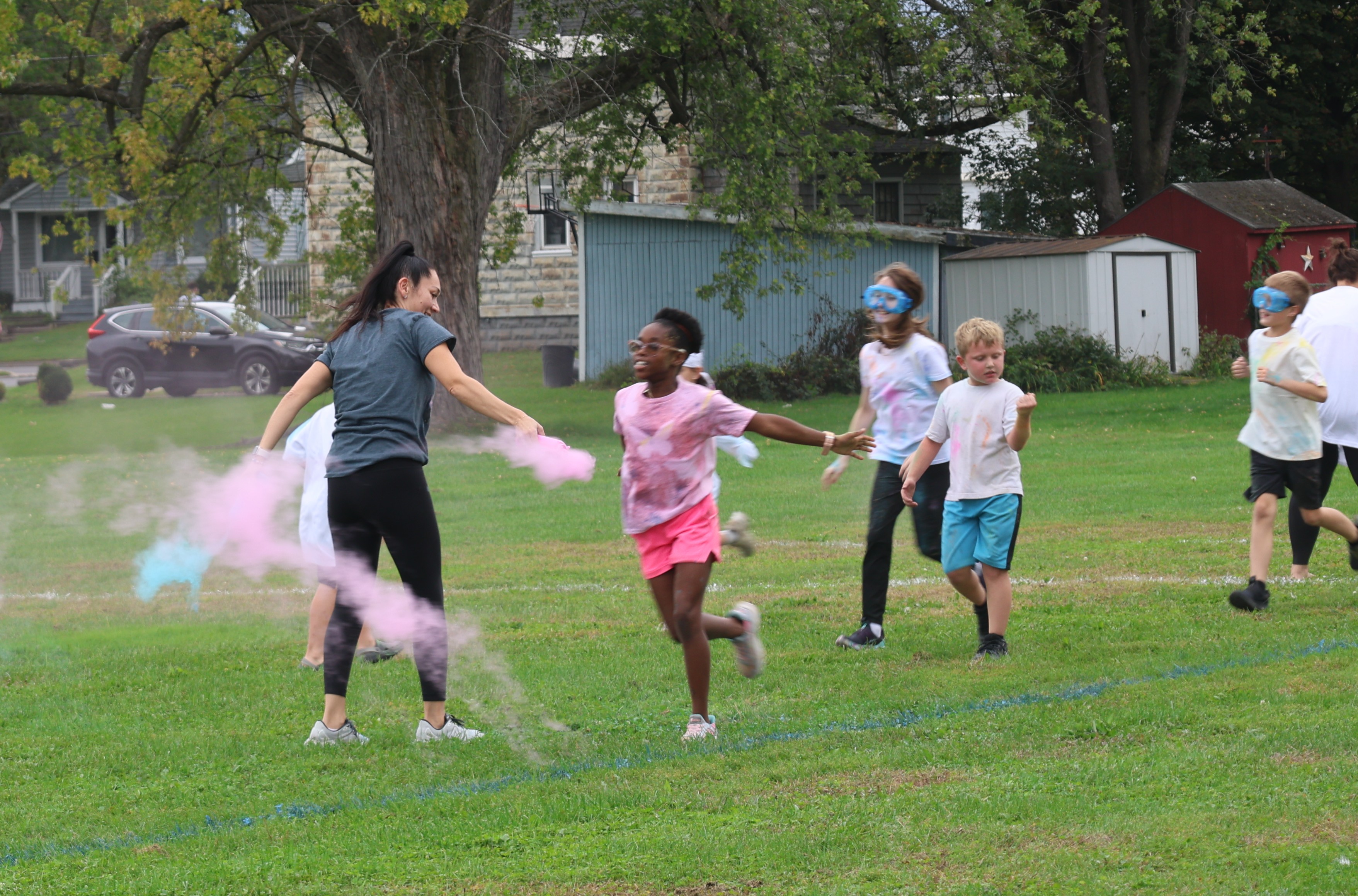 A group of children and an adult run joyfully across a grassy field during a colorful outdoor event, surrounded by clouds of pink and blue powder.