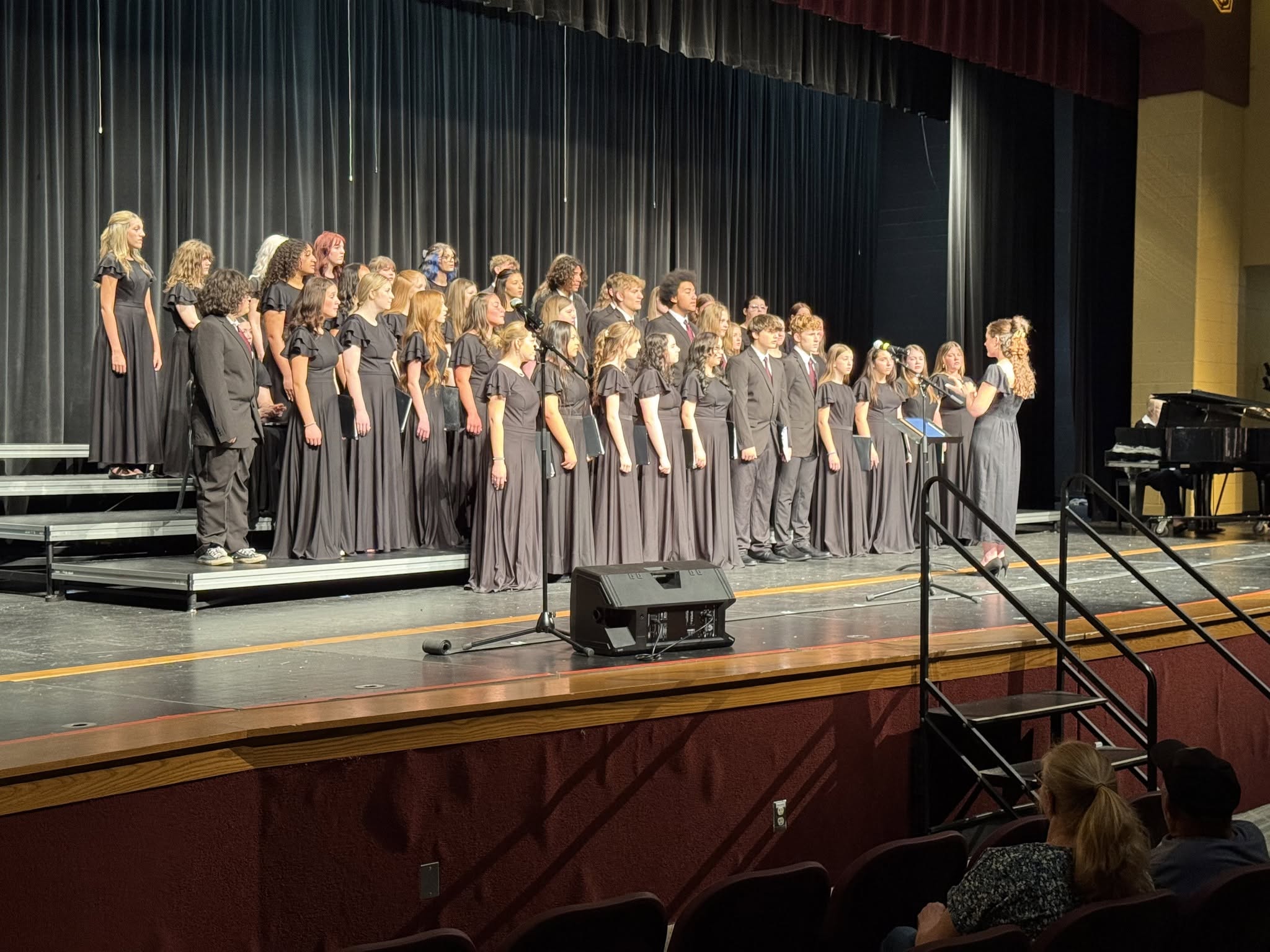 A large choir in black attire stands on risers on a stage, facing a conductor.