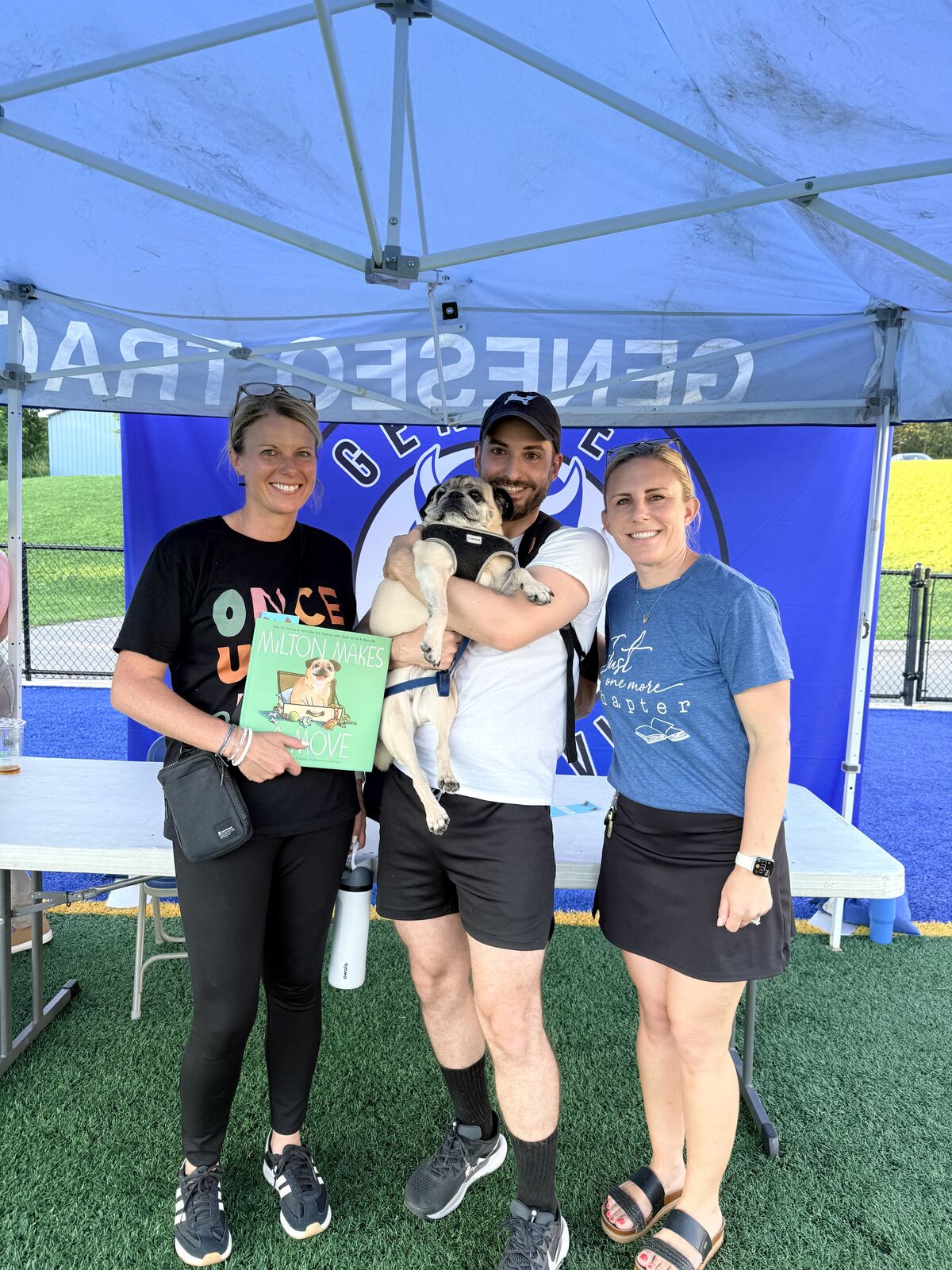 Three people and a pug smile under a blue and white tent, with a book titled 'Milton Makes a Move' visible.