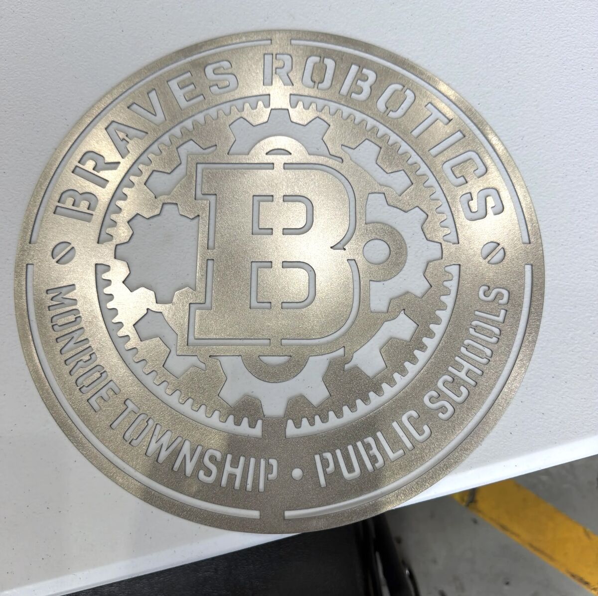 A circular metal emblem with cut-out text and gears reads 'BRAVES ROBOTICS MONROE TOWNSHIP PUBLIC SCHOOLS'.